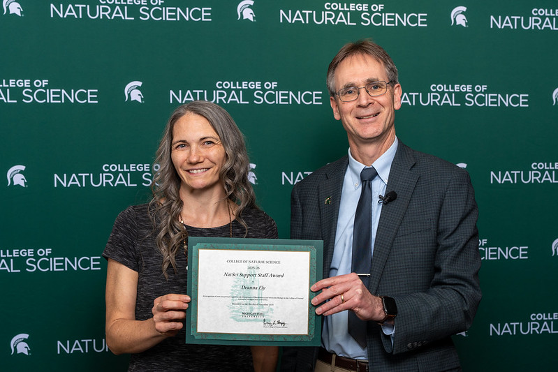 Deanna Ely receives her award from Eric Hegg in front of a green wall with white NatSci branding