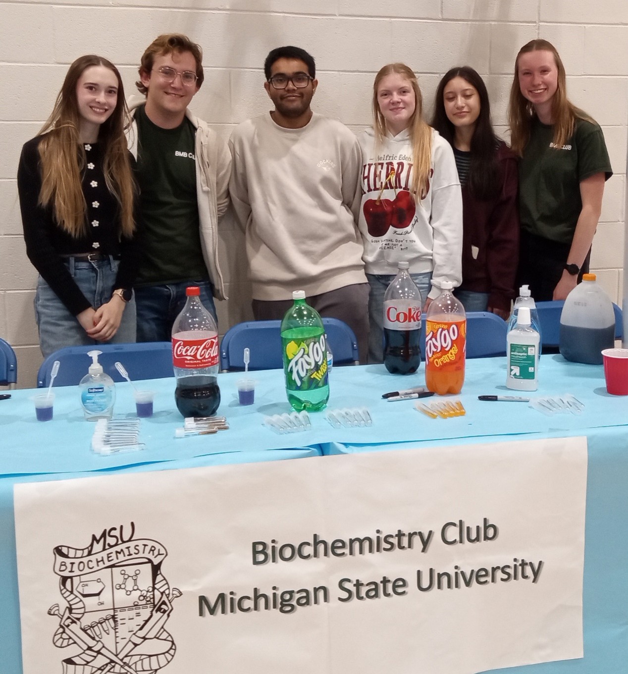 Six members of BMB Club smile near a table set up with fun chemistry experiments